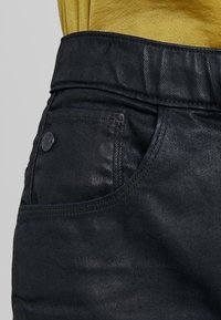 Svarta denimjeans har en framficka med en svart knapp, texturerad tyg, och en närbild på midjebandet och fickområdet.