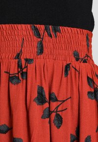 Jupe rouge avec motif de feuilles noires, dotée d'une taille smockée et d'un tissu texturé. Le design met en valeur une silhouette fluide et des détails délicats.