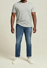 Homme portant un t-shirt à manches courtes gris clair, un jean bleu avec les revers roulés et des baskets blanches, debout devant un fond uni.