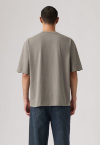 Άνδρας με κοντά σκούρα μαλλιά, φοράει μια χαλαρή γκρι κοντομάνικη μπλούζα και μπλε τζιν, γυρισμένος με την πλάτη προς ένα απλό φόντο.