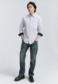Camicia a righe bianche e blu navy con bottoni e polsino blu navy solido, abbinata a jeans blu scuro e scarpe nere. Il modello posa con le mani nelle tasche.