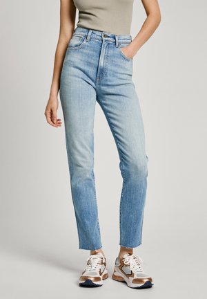 Jeans high-rise blu chiaro con vestibilità dritta, caratterizzati da un classico design a cinque tasche e orlo leggermente sfrangiato, abbinati a sneakers bianche e metalliche.