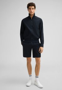 Marineblauwe ritsjack met textuurstof, gecombineerd met bijpassende shorts. Model draagt witte instappers en sokken met zwarte strepen.