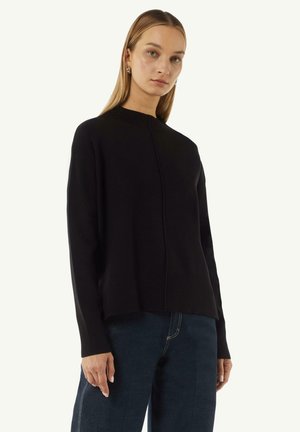 Schwarzer Strickpullover mit hohem Kragen und gerippten Kanten. Besitzt eine mittige Naht an der Vorderseite und wird mit dunkelblauen Jeans kombiniert.