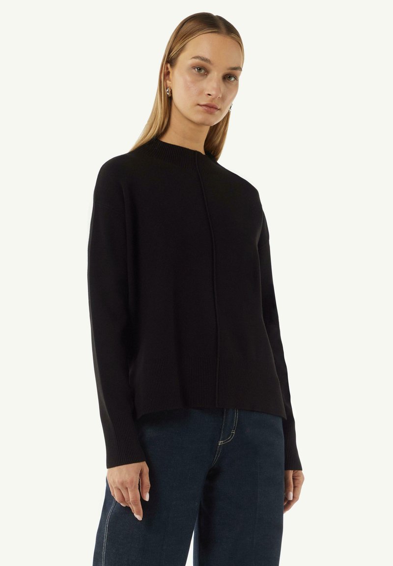 Black knitted sweater with a high neckline and ribbed edges. Features a centered seam down the front, paired with dark blue jeans.