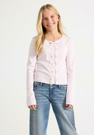 Smilende pige med langt blond hår iført en lyserød langærmet top med blonder foran og blå jeans med mørke pletter.