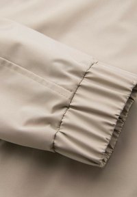 Nahaufnahme des beigen Jackenarms, der das elastische Bündchen und die glatte Stofftextur zeigt.