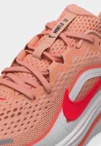 Peach atspalvio Nike bėgimo batelio uždarymas, pagamintas iš kvėpuojančio tinklelio, su rožinės spalvos raišteliais, raudonu „swoosh“ logotipu ir ant liežuvio tvirtinamoje juostoje esančiu užrašu „WmN FL 12“.
