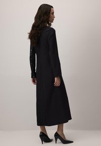 Vestido negro con mangas largas; una manga presenta lentejuelas negras. El vestido es de longitud media con una silueta recta y una abertura lateral.