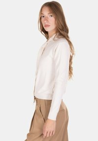 Cardigan color crema con design collo, maniche lunghe e chiusura con bottoni. Tessuto liscio, abbinato a pantaloni color tannico a vita alta.