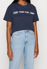 Persona vestida con una camiseta azul marino con el texto "JEANS TOMMY" y pantalones vaqueros de tiro alto azul claro, contra un fondo liso.