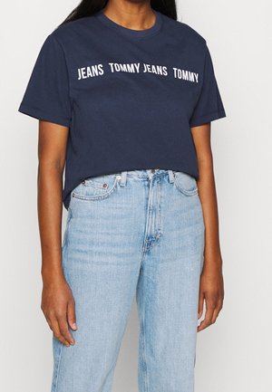 Person iført en marineblå t-shirt med teksten "JEANS TOMMY" og lyseblå højtaljede jeans foran en ensfarvet baggrund.