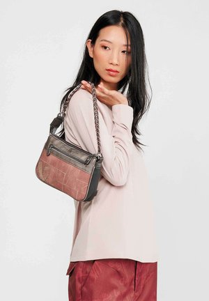 Mujer con cabello negro largo que lleva una camisa de manga larga rosa claro y pantalones rojos, sosteniendo un bolso marrón y negro con una correa trenzada sobre el hombro.