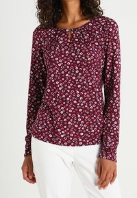 Camicia a maniche lunghe di un borgogna scuro con stampa floreale bianca e rosa. Presenta un collo a fessura e pieghe sulle spalle.