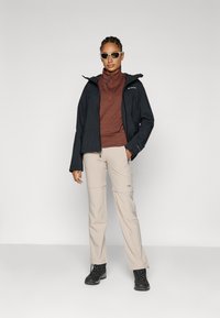 Black waterproof jacket, brown long-sleeve top, beige pants with zip-off feature. Black boots and sunglasses complete the outdoor outfit.