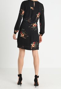 Vestido negro con bordado floral, mangas largas, un escote en forma de llave en la parte posterior y una silueta ajustada. Combinado con zapatos de tacón alto negros.