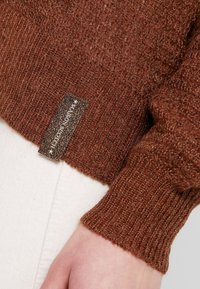 Pull en tricot marron avec une texture côtelée et des fils métalliques subtils. Présente une étiquette de marque tissée sur la couture latérale.