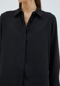 Maglietta nera a maniche lunghe con collo a camicia, dotata di chiusura con bottoni sul davanti e tessuto liscio, leggermente lucido. Design minimalista.
