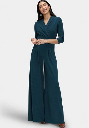Vrouw met lang donker haar draagt een teal jumpsuit met wijde pijpen en driekwartmouwen en rode open teen hakken, staand tegen een lichte achtergrond.