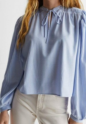 Femme portant un chemisier à rayures bleu clair avec un nœud au col et des manches longues bouffantes, assorti à un pantalon de couleur claire.