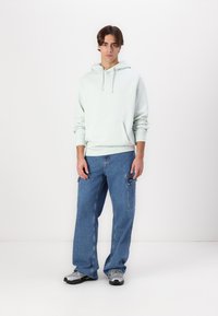 Svetlo zelena kapuca s sprednjim žepom, v kombinaciji z ohlapnimi modrimi cargo jeans hlačami, ki imajo stranske žepe. Model nosi sivo športno obutev.