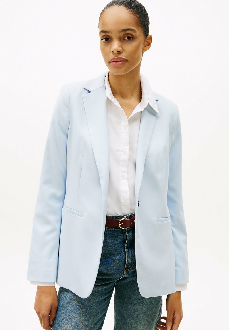 Blazer azzurro chiaro con un design strutturato, due tasche frontali e rever a crestino, abbinato a una camicia bianca con bottoni e jeans in denim.