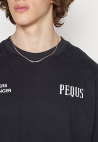 Camiseta de algodón negra con texto blanco "PEQUS" y "SADERS BANGER" en el frente. Cuello redondo con ribete y una textura suave.
