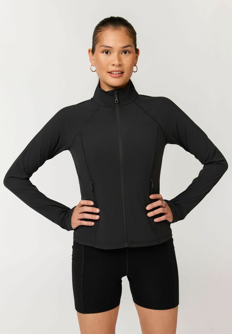 Giovane donna con i capelli raccolti che indossa una giacca nera con zip e pantaloncini da biker neri, in piedi con le mani sui fianchi contro uno sfondo semplice.