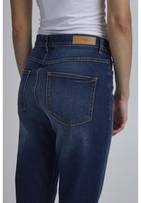 Mörkblå denimjeans med hög midja, med en läderlogg på baksidan, synlig sömnad och två bakfickor.