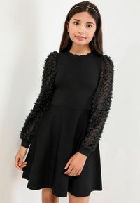 Robe noire avec de longues manches transparentes ornées d'appliqués floraux. Encolure festonnée, corsage ajusté et jupe évasée en tissu lisse.