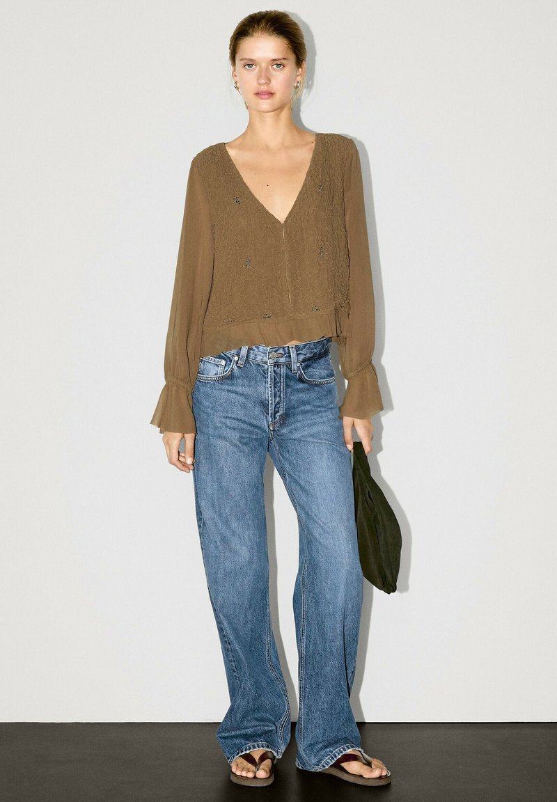 Olivegrüne, strukturierte Bluse mit V-Ausschnitt und ausgestellten Ärmeln, kombiniert mit hellblauen, weiten Jeans und braunen Sandalen.