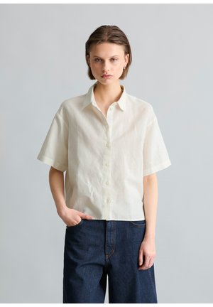 Junge Person mit kurzem Haar, die ein weißes Kurzarm-Hemd mit Knöpfen und dunkelblaue Jeans trägt, steht vor einem schlichten Hintergrund.