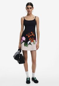 Svart figurnära klänning med blommönster i orange, rosa och grönt vid fållen. Modellen håller en svart handväska och bär svarta sneakers med gröna snören.