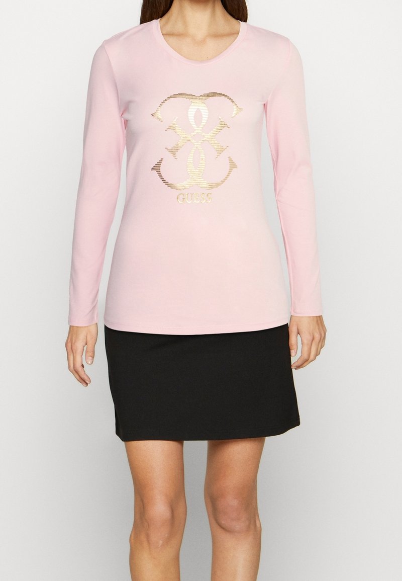 Ljust rosa långärmad skjorta med en metallisk guld logotypdesign. Kombination med en svart kjol, slät textur och avslappnad passform.