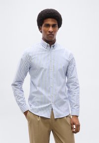 Άνδρας με αφρομαλλιά, ντυμένος με πουκάμισο με κατακόρυφες ρίγες σε ανοιχτό μπλε και άσπρο και μπεζ παντελόνι, στέκεται με το ένα χέρι στην τσέπη.