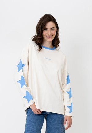 Lachende vrouw met bruin haar draagt een wit shirt met lange mouwen met blauwe sterren op de mouwen en de tekst "OH APRIL", gecombineerd met een blauwe spijkerbroek.