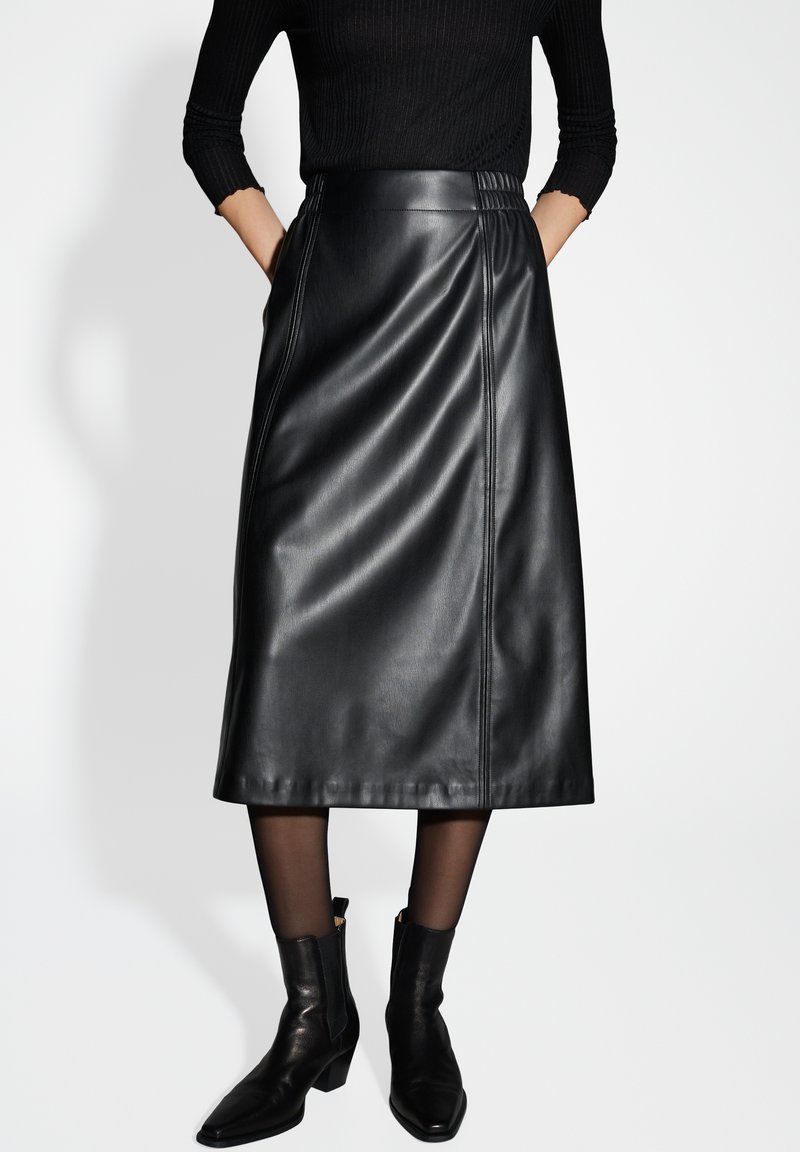 Jupe midi en cuir noir avec une texture lisse, dotée de coutures latérales. Associée à des bottes à talons noires et des collants noirs transparents.