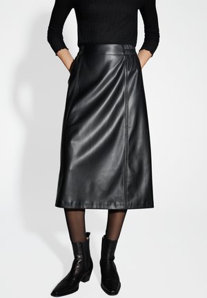 Zwarte leren midi-jurk met een gladde textuur, voorzien van zijgestikte naden. Gepaard met zwarte haklaarzen en doorschijnende zwarte panty's.