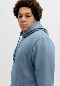 Jopica z ruto iz mehke modre tkanine, zadaj žep in vrvico za zategovanje. Model ima kratke kodraste lase in pegice, prikazan s strani.