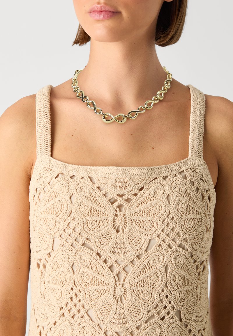 Beige, gebreide tanktop met ingewikkelde vlinderpatronen. Gouden kettinghalsketting met in elkaar grijpende lusontwerp. Soepele textuur.