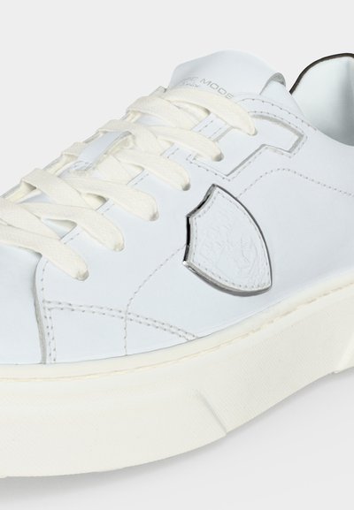 Baskets blanches avec lacets blancs, surpiqûres texturées et logo en forme de bouclier bordé d’argent sur le côté, présenté en gros plan sur un fond uni.
