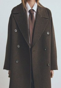 Abrigo cruzado marrón con patrón de pata de gallo, solapas anchas y botones grandes. Llevado sobre una camisa blanca y una corbata marrón.