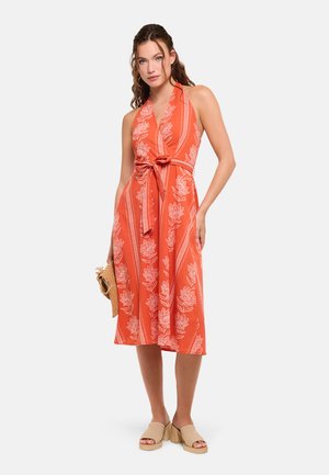Mujer con vestido sin mangas naranja con estampados florales blancos y cintura anudada, sosteniendo un clutch de paja y sandalias beige de tacón.