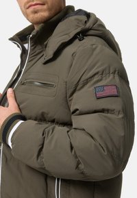 Olive green puffer jacket with hood, zipper pocket, and US flag patch on sleeve. Features ribbed cuffs and a smooth texture.