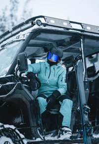 En person i en ljus turkos skidoverall med svarta detaljer och svart hjälm sitter i en svart ATV, omgiven av snö. Skidutrustning syns i närheten.