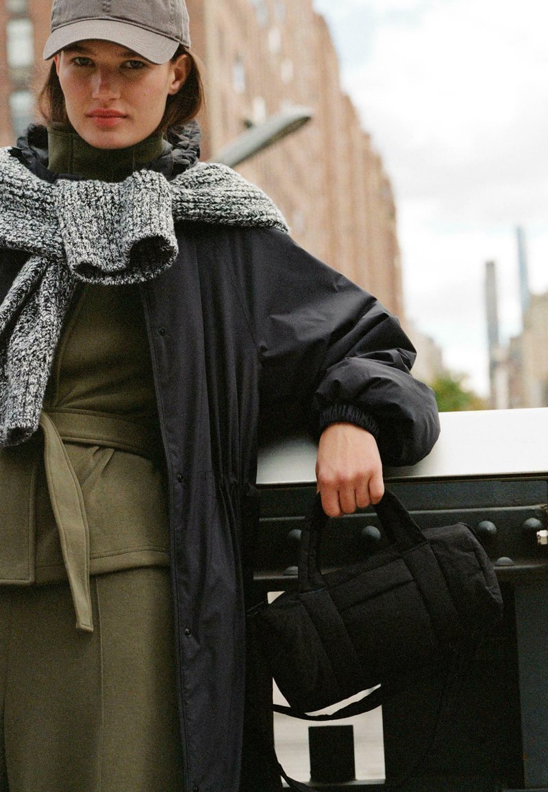Geantă neagră matlasată cu mânere scurte, purtată cu o ținută verde măsliniu și un fular tricotat gri pe un model într-un decor urban.