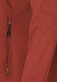 Punainen pehmeäkuorinen takki, jossa on vetoketjullinen tasku, sileä pinta ja mustat yksityiskohdat. Kevyt ja vartalonmyötäinen muotoilu.