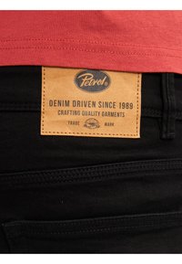 Zwarte denim jeans met een tan leren label op de tailleband waarop een logo en tekst zijn gedrukt, en twee achterzakken.