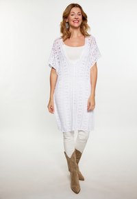 Weißes Spitzen-Tunikakleid mit V-Ausschnitt, fließenden kurzen Ärmeln und aufwendigen Strickmustern. Kombiniert mit hellen Hosen und taupefarbenen Cowboystiefeln.