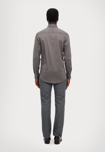 Chemise grise à manches longues avec des boutons aux poignets, un col classique et un ourlet incurvé, associée à un jean foncé et des chaussures.